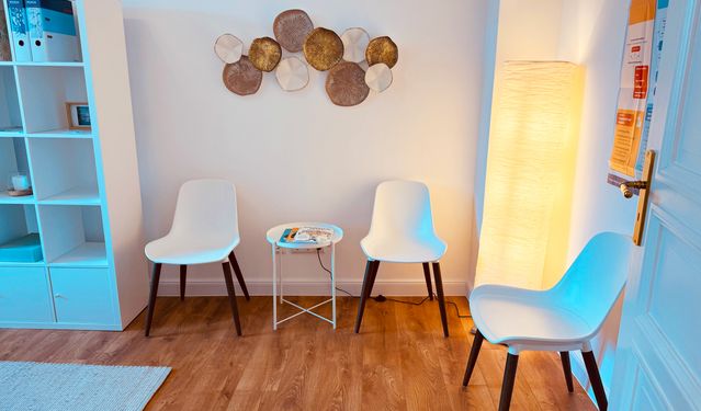 Modernes Wohnzimmer mit minimalistischer Einrichtung, bestehend aus einem grünen Sofa mit neutralen Zierkissen, einem runden Holzcouchtisch mit einer Keramikvase, einem beigefarbenen Sessel und Zimmerpflanzen. Natürliches Licht fällt durch große Fenster mit taupefarbenen Vorhängen und beleuchtet den beigefarbenen Teppich auf dem Holzboden.