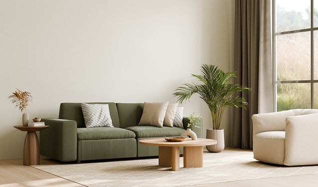 Modernes Wohnzimmer mit minimalistischer Einrichtung, bestehend aus einem grünen Sofa mit neutralen Zierkissen, einem runden Holzcouchtisch mit einer Keramikvase, einem beigefarbenen Sessel und Zimmerpflanzen. Natürliches Licht fällt durch große Fenster mit taupefarbenen Vorhängen und beleuchtet den beigefarbenen Teppich auf dem Holzboden.