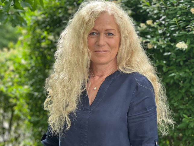 M.Sc. Psych. Katrin Bonato-Ehrenthal mit langen, lockigen, blonden Haaren in dunkelblauer Bluse, stehend vor einem blühenden grünen Hintergrund im Freien – mit freundlichem und professionellem Ausdruck.